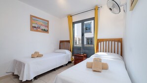 Ein Hotelzimmer mit zwei Einzelbetten, einem Nachttisch, einem Wandventilator, einem Fenster mit Vorhängen und einem Bild an der Wand.