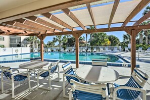 A covered pool area with lounge chairs and tables, a clear blue pool, and a view of palm trees and buildings in the background. - Walk to St Augustine Beach! Resort-Style Condo (St. Augustine)