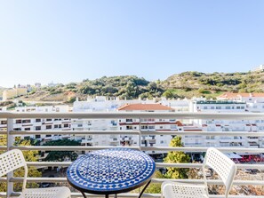 Ein Balkon mit rundem Tisch und zwei Stühlen, mit Blick auf ein Wohngebiet mit weißen Gebäuden und einer Hügelkulisse im Hintergrund.