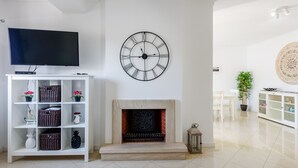 A living room with a flat-screen TV, a decorative wall clock, a fireplace, and a white shelving unit with various items. - Apartment  by Interhome (Albufeira)
