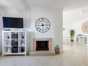 Ein Wohnzimmer mit Flachbildfernseher, einer Wanduhr, einem Kamin und einer weißen Regalanlage mit diversen Gegenständen.