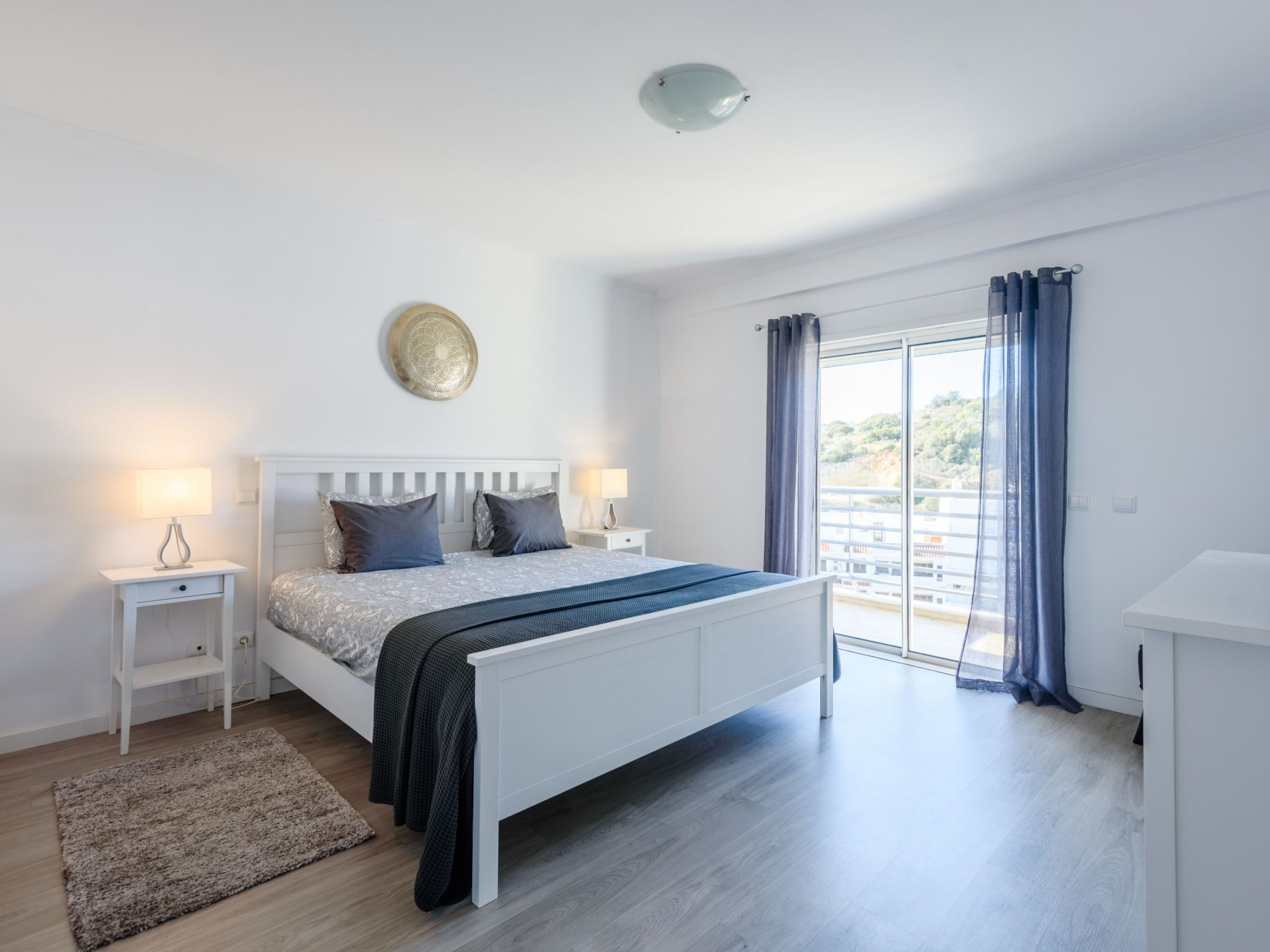 Un dormitorio moderno con una cama grande, mesitas de noche, una lámpara de pared y vistas a través de una ventana con cortinas.