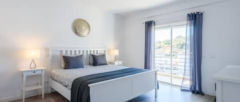 Un dormitorio moderno con una cama grande, mesitas de noche, una lámpara de pared y vistas a través de una ventana con cortinas.