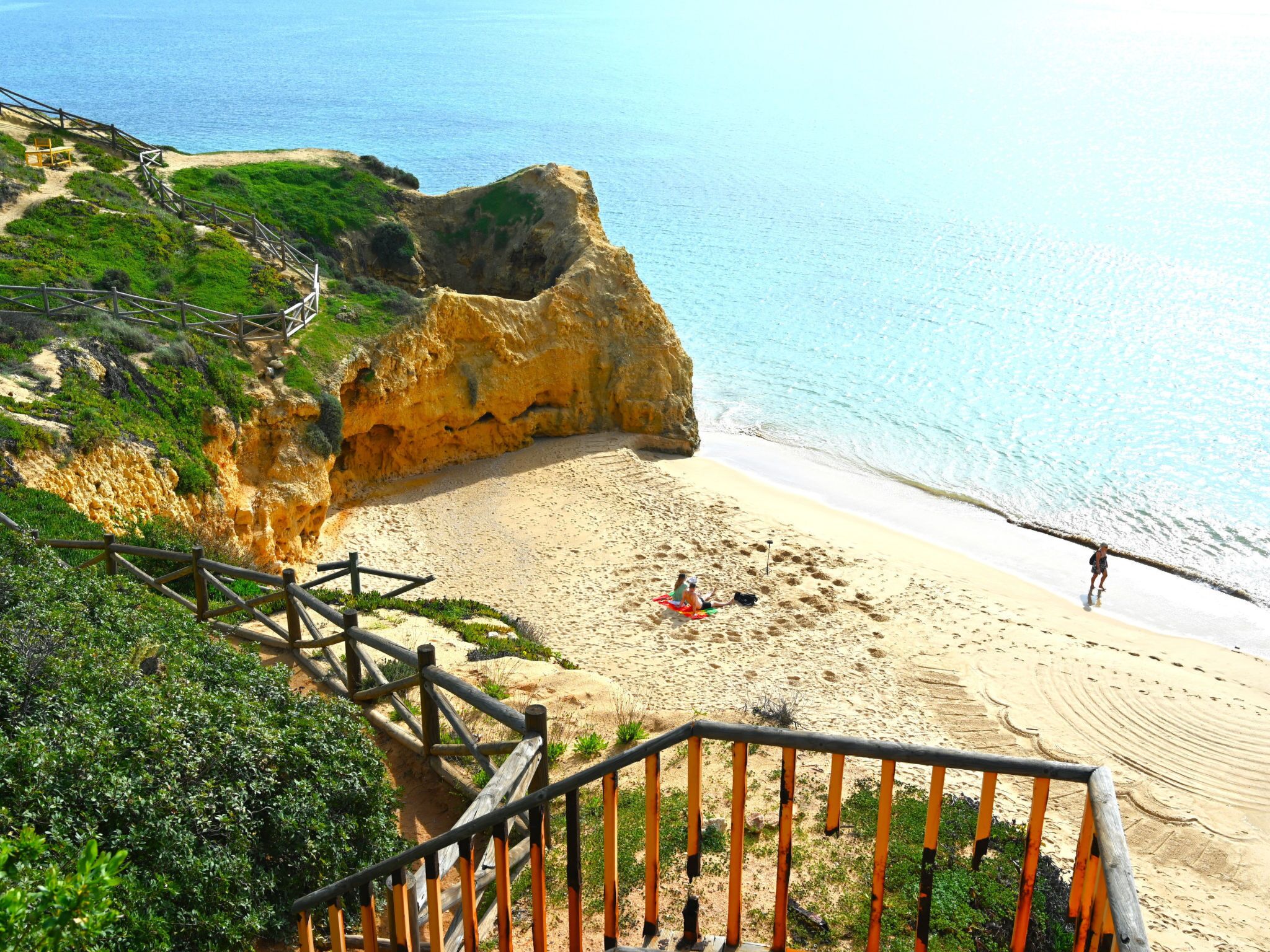 A coastal beach with a wooden staircase leading down to the sand, a clear view of the ocean, and a person walking along the beach.