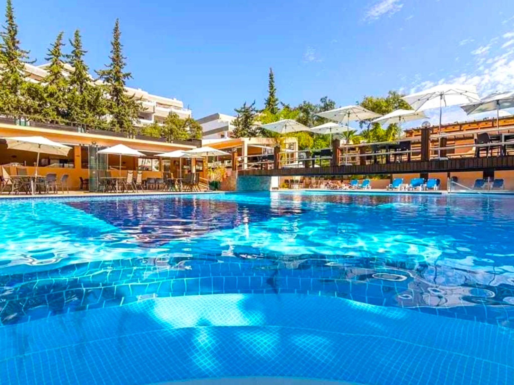 A swimming pool with clear blue water, surrounded by lounge chairs and umbrellas, and a building with multiple balconies in the background.