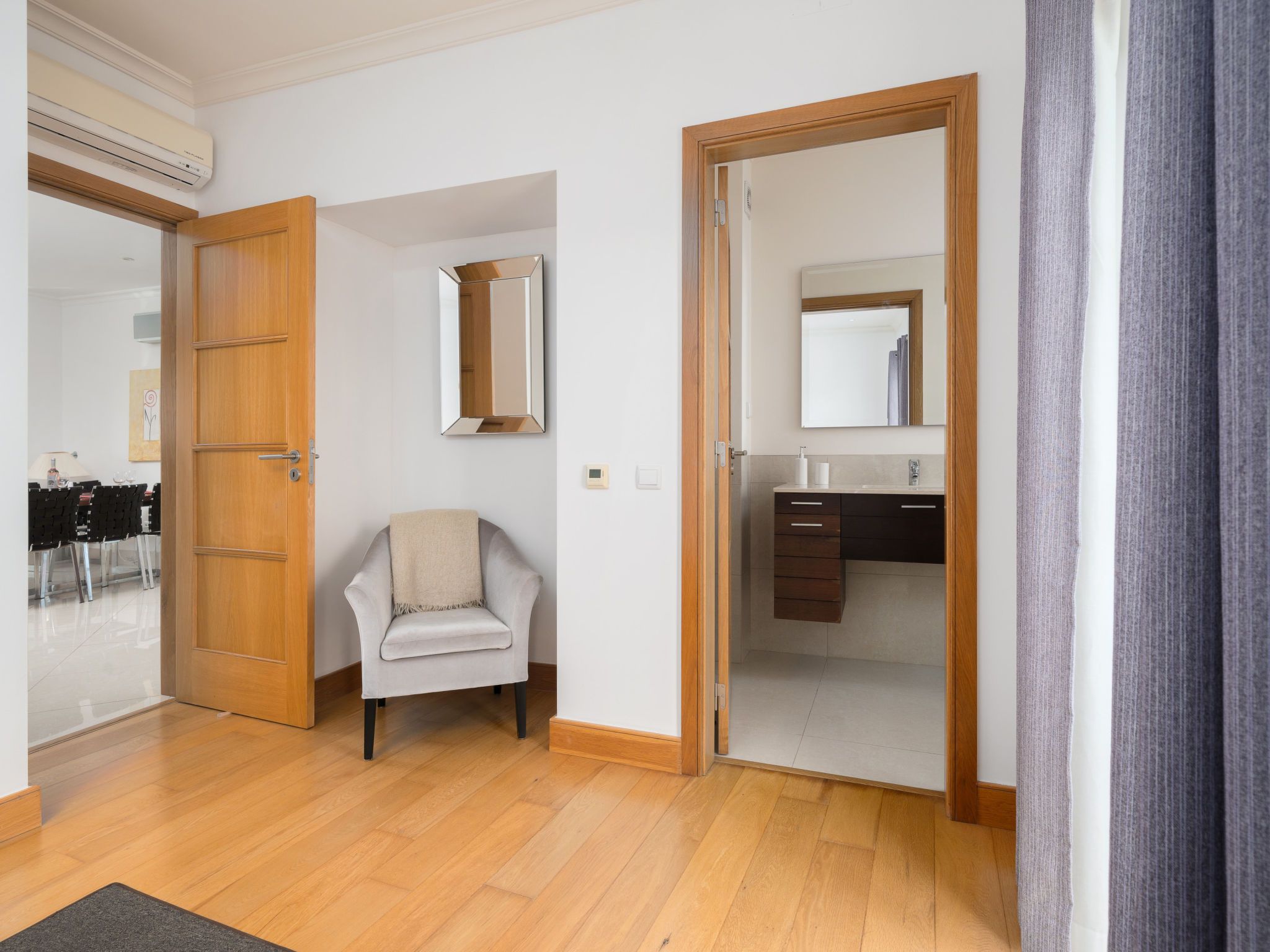 Een hotelkamer met een houten vloer, een stoel, een bed, een badkamer met een spiegel en een deur naar een andere kamer.