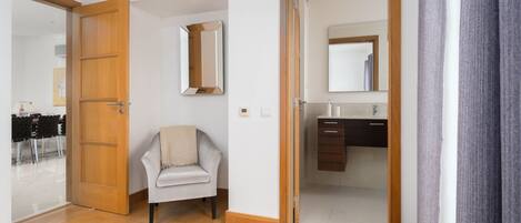 Een hotelkamer met een houten vloer, een stoel, een bed, een badkamer met een spiegel en een deur naar een andere kamer.