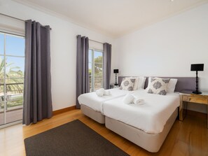 A hotel room with a large bed, two bedside tables, a desk, and a view of palm trees through the window. - Apartment  by Interhome (Quarteira)