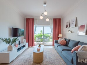 A modern living room with a flat-screen TV, a sofa, a coffee table, and a view of a garden through large windows. - Apartment  by Interhome (Lagos)