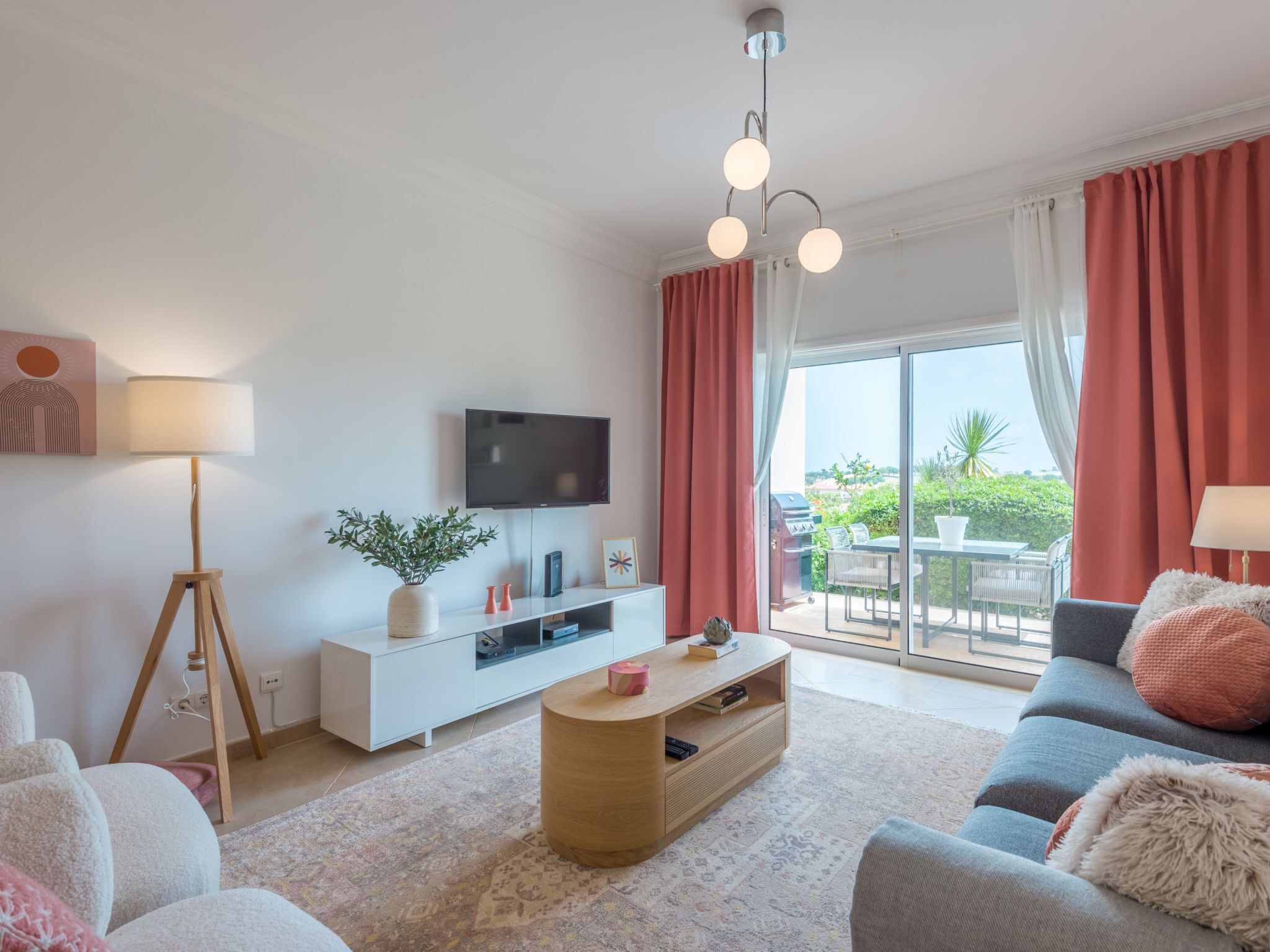 Ein modernes Wohnzimmer mit Flachbildfernseher, Holztisch, einer Couch und einem großen Fenster mit Vorhängen.