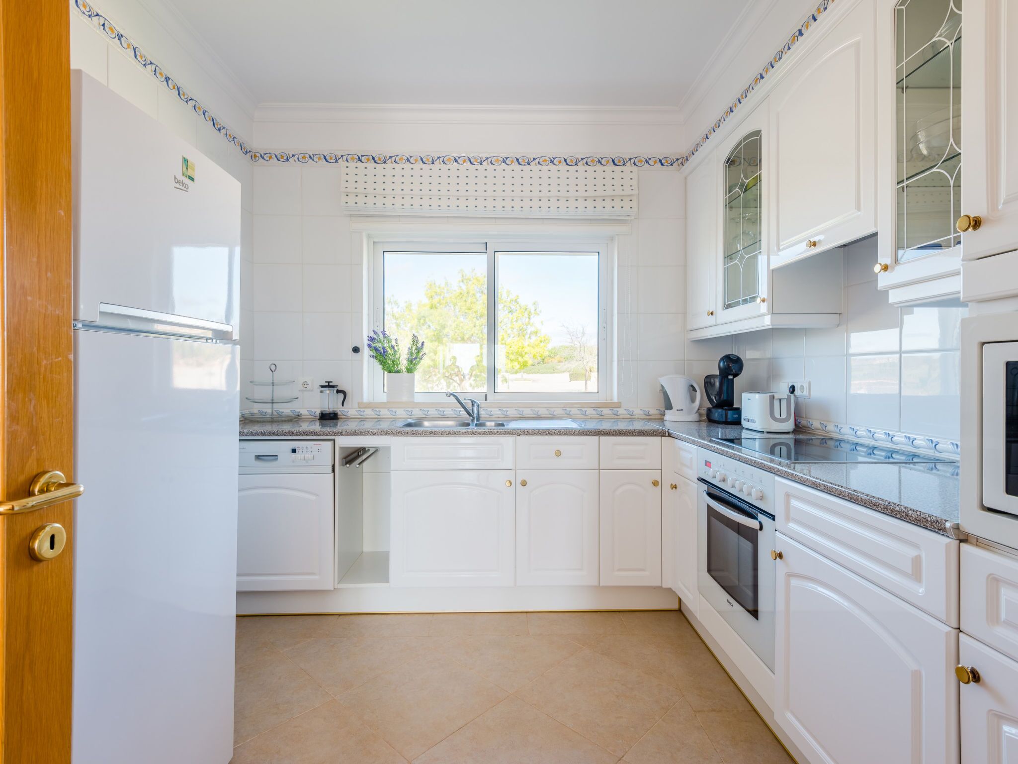 A kitchen with white cabinets, a built-in oven, a dishwasher, and a window with a view of greenery.