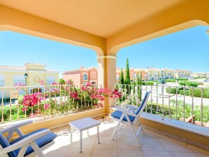 Ein Balkon mit zwei Lounge-Stühlen und einem kleinen Tisch, mit Blick auf ein Wohngebiet mit bunten Gebäuden und üppiger Vegetation.