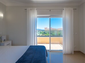 A bedroom with a bed, a nightstand, a window with curtains, and a view of a balcony and buildings. - Vacation home  by Interhome (Lagos)