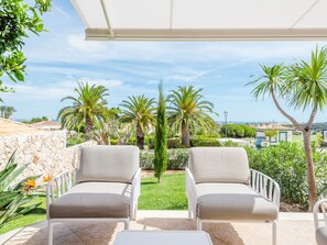 Eine Terrasse mit zwei weißen Stühlen und einem Tisch, mit Blick auf einen Garten mit Palmen und Meer.