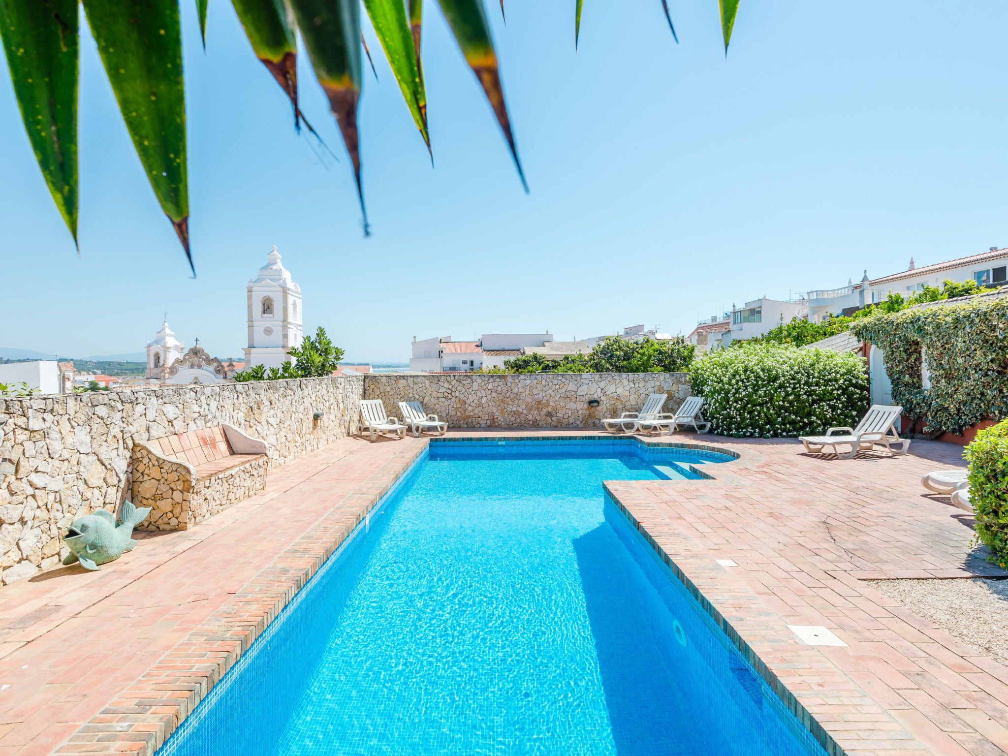 A swimming pool with a stone wall and lounge chairs, a view of a church, and buildings in the background.