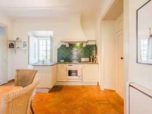 Une cuisine avec des meubles blancs, un pare-feu en carrelage, un four encastré et une chaise en osier.