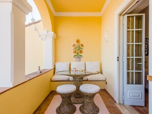 Un petit balcon fermé, agrémenté d’une table ronde en verre, de deux tabourets blancs avec coussins, d’un canapé blanc et d’un vase décoratif rempli de tournesols.