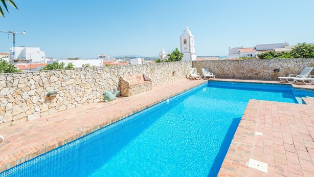 A clear blue swimming pool with stone walls and a stone bench, surrounded by a stone wall and a view of buildings and a church in the background.