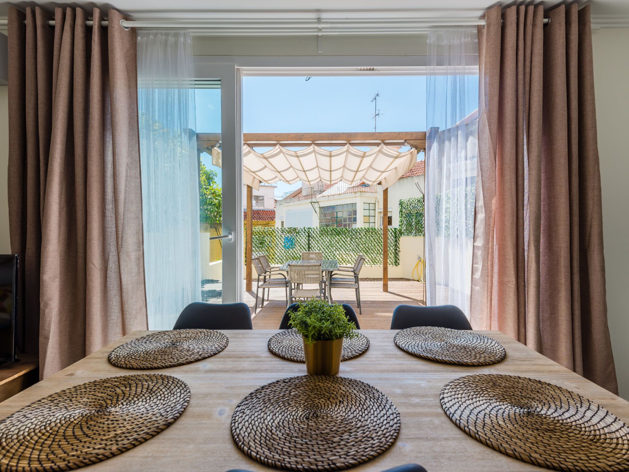 Un coin repas avec une table dressée pour six personnes, une plante en pot et une vue sur une terrasse extérieure avec une table et des chaises.
