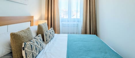Et hotelværelse med seng, puder, sengegavl, et vindue med gardiner og et billede på væggen.