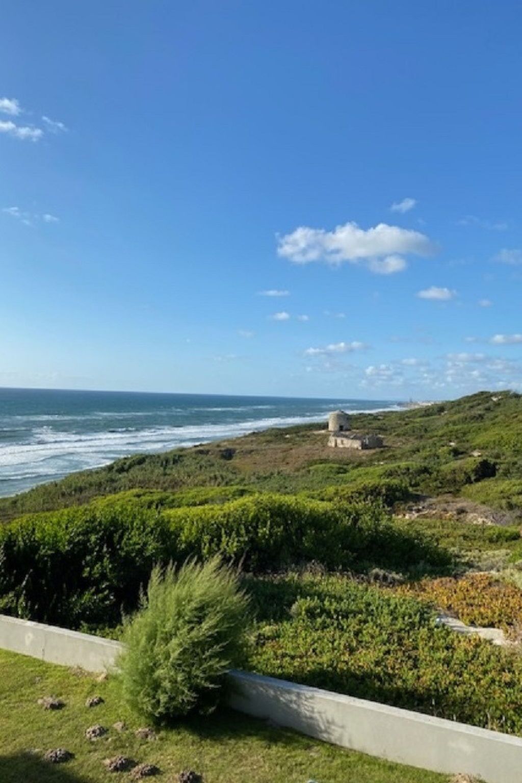 A coastal landscape with a grassy area, a small building, and the ocean in the background.