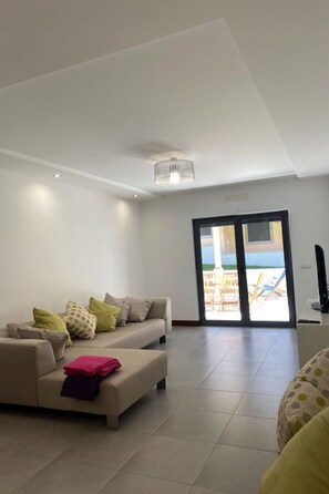 A modern living room with a sofa, a coffee table, and a television. There is a sliding glass door leading to an outdoor area. - Apartment  by Interhome (Alcobaça)