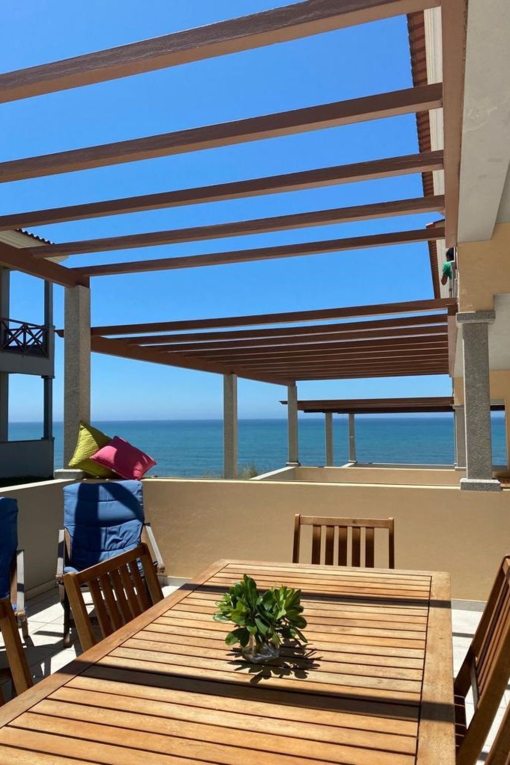 A wooden table with chairs and a potted plant, set under a pergola with a view of the sea.