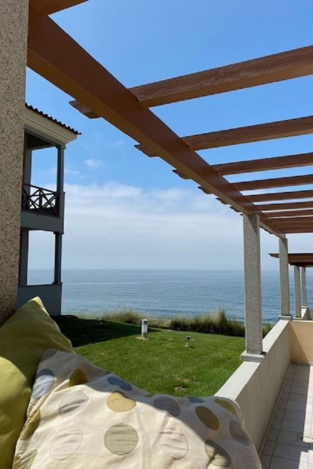 A covered outdoor area with a view of the sea, a building with a balcony, and a grassy area.