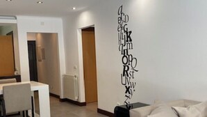 Een moderne woonkamer met een bank, een bijzettafeltje en een muur met letters van het alfabet. Op de achtergrond is een eethoek met een tafel en stoelen te zien.