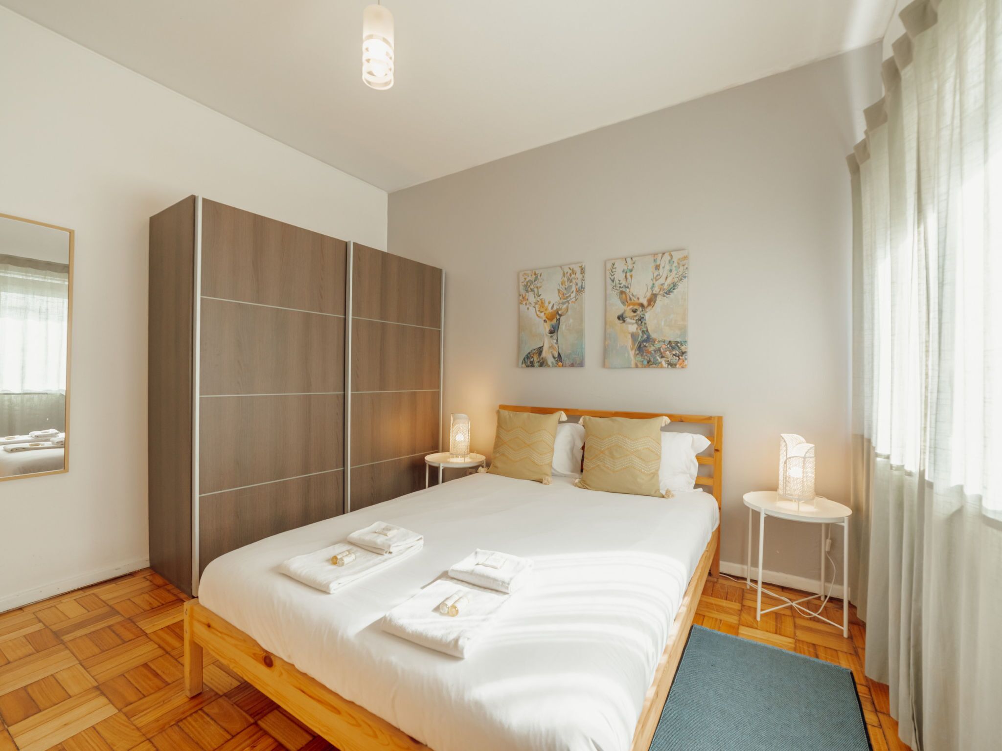 A modern bedroom with a large bed, a wardrobe, two paintings, bedside lamps, and a view of the outdoors.