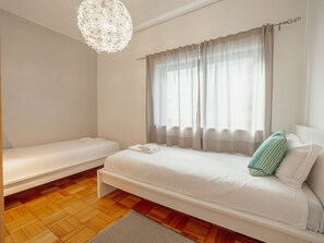 A bedroom with two single beds, a large window with curtains, a chandelier, and a wooden parquet floor.