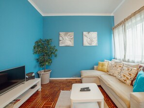 A living room with a blue wall, a sofa with patterned cushions, a TV, a coffee table, and a potted plant.
