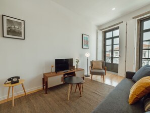 Ein Wohnzimmer mit einer Couch, einem Fernseher, einem kleinen Tisch, einem Sessel und einer Stehlampe.