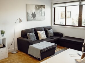 A modern living room with a black sofa, a side table, a floor lamp, and a window with a view of trees and buildings. - DOWNTOWN STYLISH APARTMENT & GARAGE by VACATIONY by Interhome (Porto)