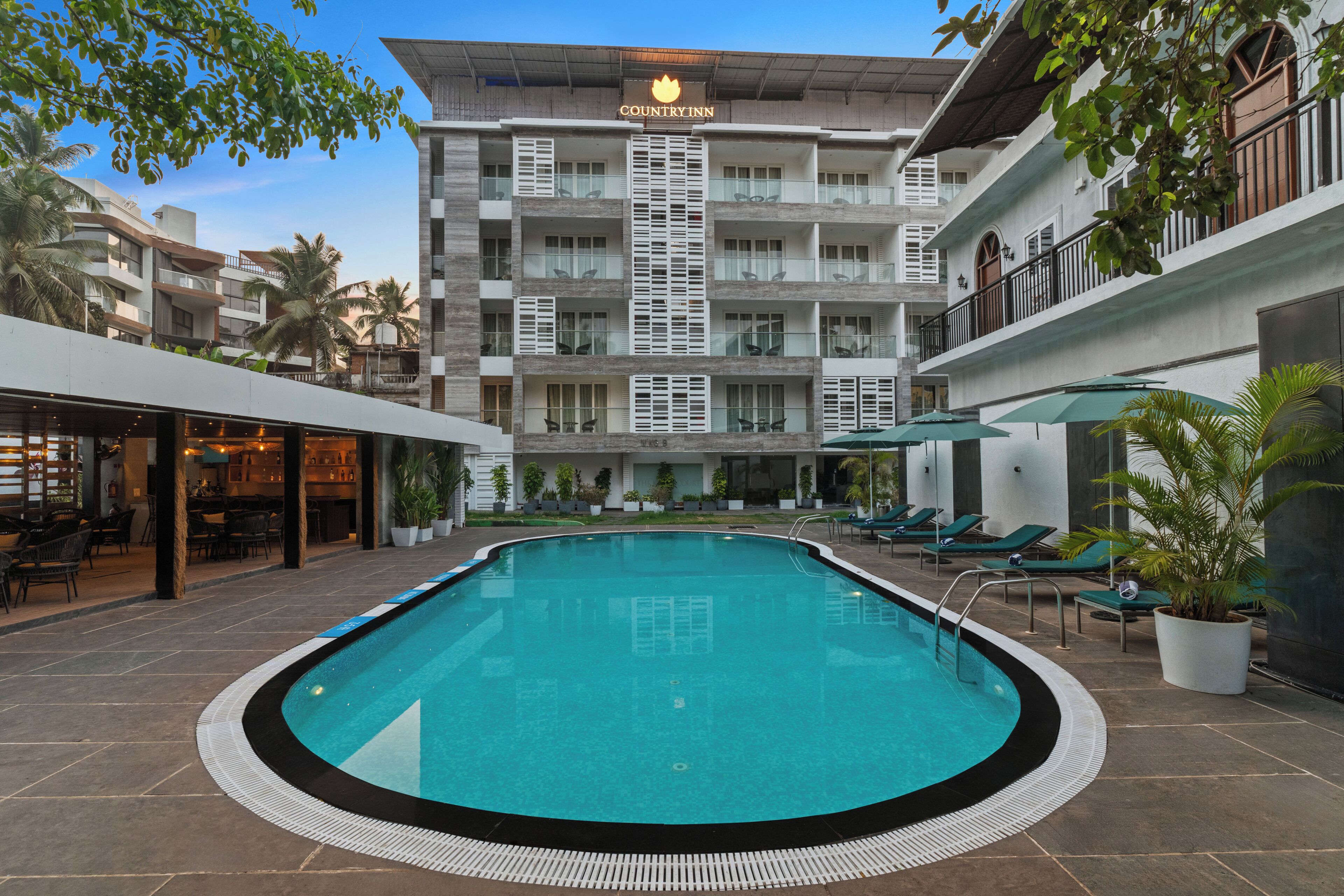 A pool area with lounge chairs, a restaurant, and a multi-story building with balconies.