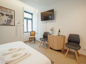 Ein Hotelzimmer mit einem Bett, zwei Stühlen, einem kleinen Holztisch, einem an der Wand befestigten Fernseher und einem Fenster mit Vorhängen.