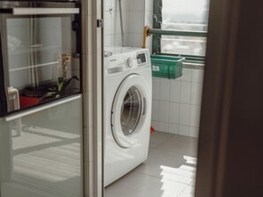 Eine Waschmaschine in Weiß ist von vorne zu sehen. Im Waschraum befindet sich ein Fenster und ein grüner Wäschekorb an der Wand.