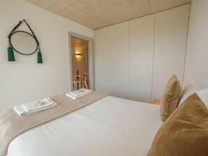 Un lit bien fait, avec un matelas blanc, une couverture beige et deux oreillers. On aperçoit, au fond, une petite table avec une chaise, ainsi qu’une armoire dotée d’un miroir et d’une décoration en forme de pompon vert.