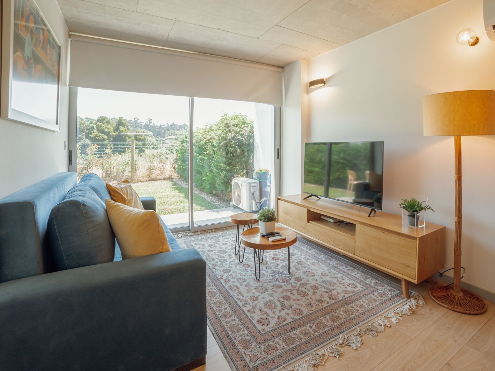A modern living room with a sofa, TV, and a view of a garden through large windows.