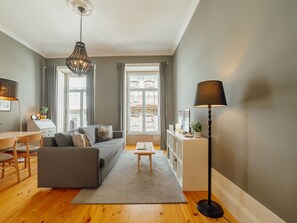 Una sala de estar moderna con un sofá gris, suelo de madera, un área de comedor con mesa y sillas, una mesita de centro, una lámpara de pie y un amplio ventanal con cortinas.