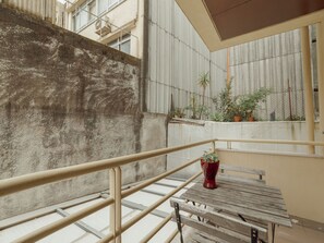 Ein Balkon mit einer Betonwand, einem Metallgeländer und einer hölzernen Bank. Auf dem Geländer stehen Blumentöpfe und auf der Bank eine rote Vase.