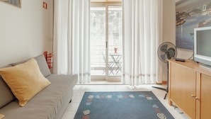 Ein Wohnzimmer mit einer Couch, einem Fernseher, einem Ventilator und Blick auf einen Balkon mit Gartenmöbeln.
