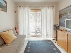 Ein Wohnzimmer mit einer Couch, einem Fernseher, einem Ventilator und Blick auf einen Balkon mit Gartenmöbeln.