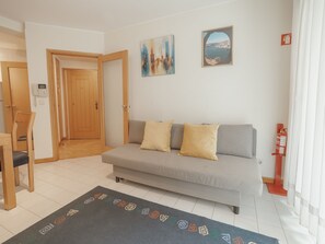 Una sala de estar con un sofá, dos cojines, una mesita de centro y una alfombra. Hay una puerta que conduce a otra habitación y un extintor de incendios montado en la pared.