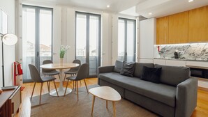 En moderne stue med en grå sofa, et rundt spisebord med stole, og et køkkenområde med træskabe og en marmorbeklædt bagvæg.