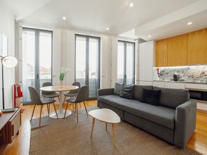 Ein modernes Wohnzimmer mit einem grauen Sofa, einem runden Esstisch mit Stühlen und einer Küchenzeile mit Holzschränken und einer Marmor-Spritzschutzleiste.