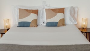 En seng med hvid madras, to puder med abstrakte mønstre og to sengeborde med lampe og flettet design.