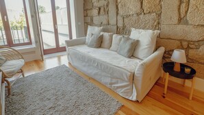 Un saló modern amb una paret de pedra, un sofà blanc amb coixins, una catifa gris i una petita taula auxiliar amb una làmpada.