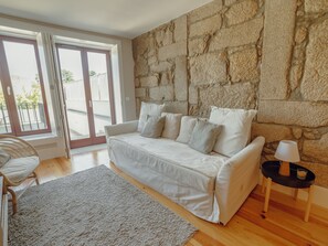 A modern living room with a stone wall, a white sofa with cushions, a grey rug, and a small side table with a lamp. - Apartment  by Interhome (Porto)