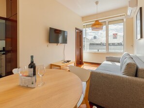 Ein modernes Wohnzimmer mit einer Couch, einem Esstisch mit Weingläsern und einer Weinflasche, einem an der Wand montierten Fernseher und einem großen Fenster mit Blick.